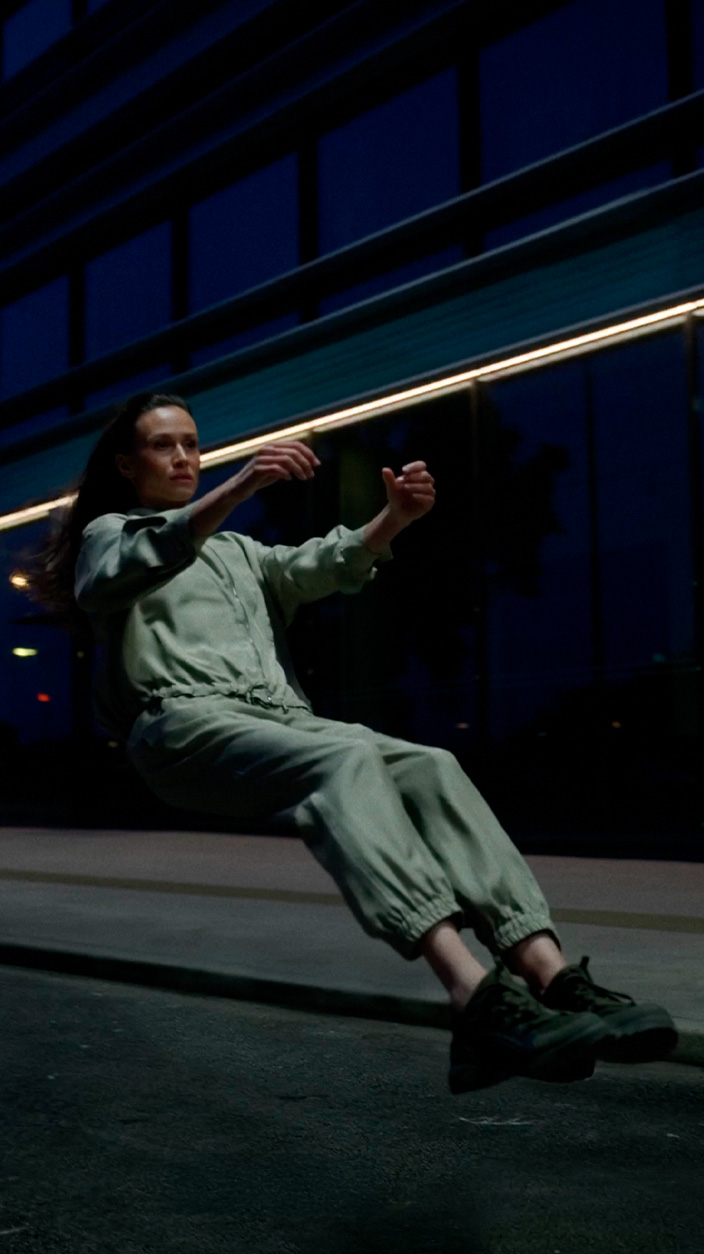 A woman in a light green jumpsuit and dark sneakers appearing to float in a seated position on a city street at night, with a modern glass building in the background.